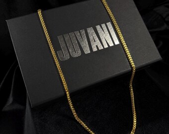 Könnte beinhalten: Eine goldfarbene Halskette liegt auf einer schwarzen Schachtel mit dem Wort "JUVANI" in Silber. Die Halskette hat ein gewebtes Design. Die Schachtel liegt auf einem schwarzen Stoffhintergrund.