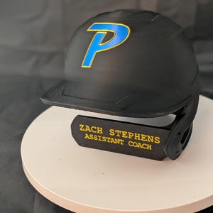 May include: A matte black baseball batting helmet featuring a blue letter P with a gold outline on the front. A black chin guard is attached to the side, displaying the yellow text ZACH STEPHENS ASSISTANT COACH in a bold, sans-serif font.