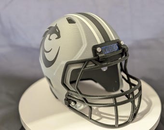Puede incluir: Un casco de fútbol americano plateado con rayas negras y una máscara facial negra. El casco presenta un logotipo de gato montés negro y la palabra "WILDCATS" en azul.