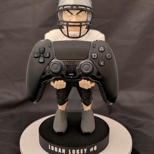May include: A figurine of a football player wearing a silver helmet and white jersey, holding a black video game controller. The figure stands on a round black base with the white text LOGAN LOKEY #6 printed on the front of the base.