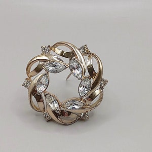 May include: A gold-toned brooch with a circular design featuring clear rhinestones set in a swirling pattern.