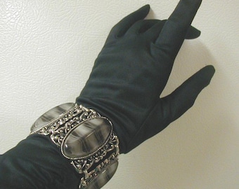 May include: A silver and black wide bracelet with an ornate design and a large oval-shaped stone. The bracelet is worn on a black glove.