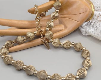 May include: Gold-coloured necklace with textured, knotted beads and smaller, round beads. The necklace has a delicate chain and a pointed pendant. The jewellery is displayed on a wooden hand.