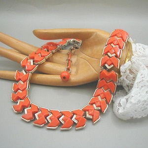 May include: An orange vintage necklace with a geometric design. The necklace features interlocking orange segments with silver-colored metal accents. A matching orange bead hangs from the clasp. The necklace is displayed on a wooden hand.