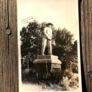May include: Black and white photograph of a statue of a man in a cowboy hat standing on a pedestal. The statue is in front of a group of trees. The text "Boot Hill Dodge City KANS" is written in the top left corner of the image.