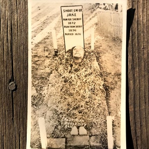 May include: A black and white photograph of a grave marker with the text "Shoot 'em Up Jake Run for Sheriff 1872 Run from Sheriff 1876 Buried 1876".