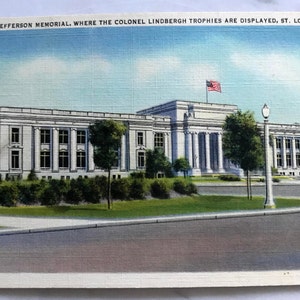 Może przedstawiać: Winietkowa pocztówka przedstawiająca Jefferson Memorial w St. Louis w stanie Missouri. Budynek jest biały z dużą, centralną kopułą i rzędem kolumn. Amerykańska flaga powiewa na maszcie na dachu. Tekst pocztówki brzmi "Jefferson Memorial, where the Colonel Lindbergh Trophies are displayed, St. Louis, Mo."