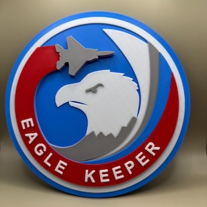 May include: A blue, white, and red circular sign with the words "Eagle Keeper" in white lettering on a red background. A gray eagle head with a gray fighter jet flying over it is in the center of the sign.
