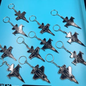 May include: A collection of twelve metal keychains, each featuring a black and silver enamel design of a fighter jet. The keychains are attached to circular silver split rings by short metal chains, arranged in a scattered pattern.