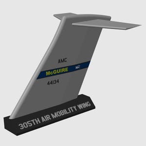 Può includere: Modello grigio di un'ala di coda di aereo, con un'ala orizzontale. L'ala di coda presenta il testo "AMC", "McGuire" e "44134" su una striscia blu. La base recita "305TH AIR MOBILITY WING".