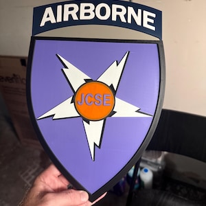 May include: A shield-shaped wall plaque featuring a purple background with a white five-pointed star and lightning bolt design. An orange circular center displays the text JCSE in purple. A dark blue tab at the top bears the white text AIRBORNE.