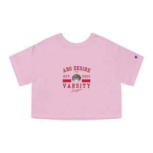 May include: A light pink crop top with a crew neck and short sleeves. The front features a red and white graphic with the text "ABO DESIRE EST. 2025 VARSITY League" and a cartoon face. A small Champion logo is on the left sleeve.