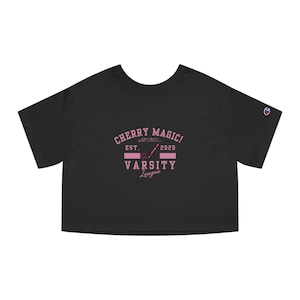 Op de afbeelding: Zwart getailleerd T-shirt met een roze afbeelding met de tekst "CHERRY MAGIC! EST. 2020 VARSITY League" met een roze honkbalknuppel en een hart.