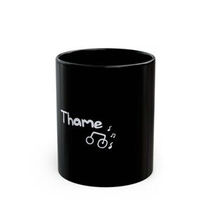 May include: Black ceramic mug with the word "Thame" in white script, accompanied by a white graphic of musical notes and a bicycle. The mug is cylindrical and stands upright.
