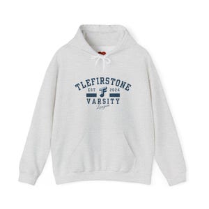 Op de afbeelding: Een lichtgrijze hoodie met een voorzak en een capuchon met trekkoord. De voorkant toont de tekst "TLEFIRSTONE VARSITY League" in marineblauw, met "EST 2024" erboven. Een casual, comfortabel sweatshirt.