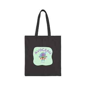May include: Black tote bag featuring a colorful graphic of a cartoon character in a life preserver with the word "AVOCEAN" in a playful font. The design is on a light green background. The bag has two black handles.