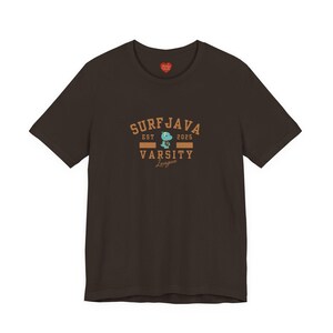 Könnte beinhalten: Dunkelbraunes T-Shirt mit dem Text "SURFJAVA VARSITY League" in Beige und einer Cartoon-Dinosaurier-Grafik. Der Text enthält "EST 2025". Ein kleines rotes Herz ist am Ausschnitt zu sehen.