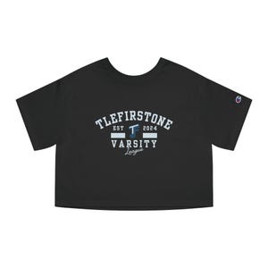 Op de afbeelding: Zwart cropped T-shirt met de tekst "TLEFIRSTONE EST 2024 VARSITY League" in wit en lichtblauw. Het Champion-logo is zichtbaar op de mouw. Het shirt heeft een ronde hals en korte mouwen.
