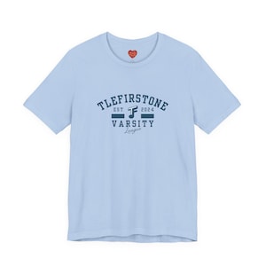 Op de afbeelding: Lichtblauw T-shirt met een marineblauwe opdruk met de tekst "TLEFIRSTONE EST 2024 VARSITY League". Een klein rood hartje staat bovenaan. Het shirt heeft korte mouwen en een ronde hals.
