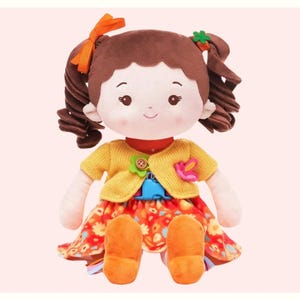 May include: A plush doll with brown curly pigtails, orange bows, and a smiling face. The doll wears a yellow sweater, a floral dress, and orange shoes. The doll has a red collar and is decorated with flower accents.