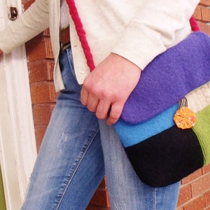 Może przedstawiać: Kobieta ma na sobie biały sweter i niebieskie dżinsy. Niesie wielokolorową torbę filcową z jasnym pomarańczowym akcentem kwiatowym. Torba ma fioletowy, niebieski, czarny i zielony wzór.
