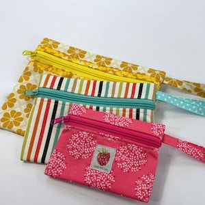 May include: Three zippered fabric pouches with wrist straps. The largest has a yellow and white floral pattern, the middle has vertical stripes in teal, black, and white, and the smallest is pink with white floral motifs and a raspberry label.