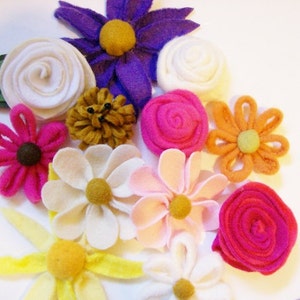 May include: A collection of felt flower embellishments in various colors, including white, pink, purple, orange, and yellow. The flowers are made of felt and have a variety of shapes and sizes.