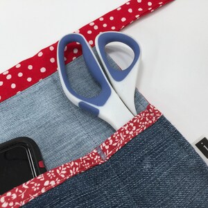 May include: A denim apron with a red and white polka dot trim, a floral trim, and a pocket that holds a pair of scissors and a smartphone. The apron has a black label with the letter "J" on it.