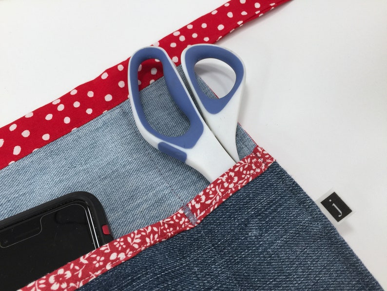 May include: A denim apron with a red and white polka dot trim, a floral trim, and a pocket that holds a pair of scissors and a smartphone. The apron has a black label with the letter "J" on it.