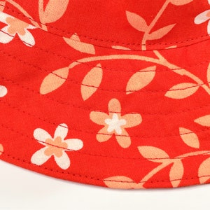 May include: Red bucket hat with a floral pattern featuring white flowers with peach centers and peach leaves on a red background.