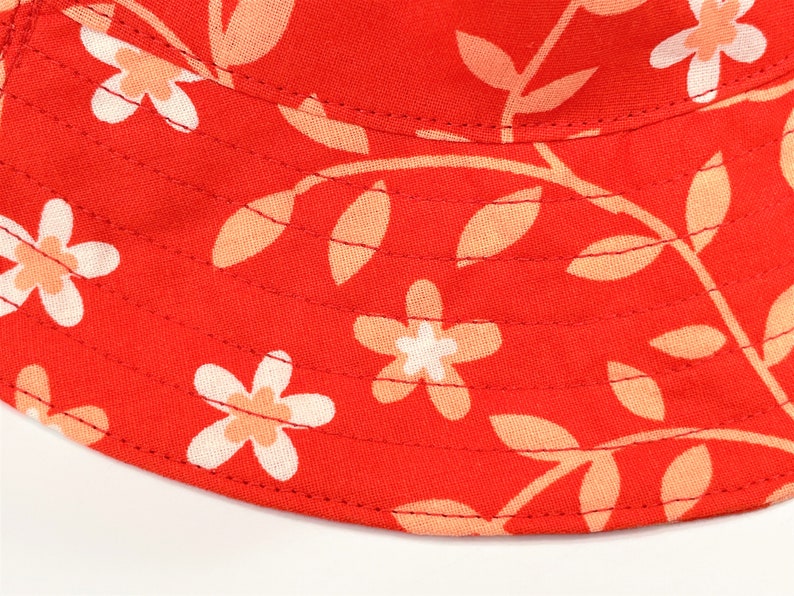 May include: Red bucket hat with a floral pattern featuring white flowers with peach centers and peach leaves on a red background.