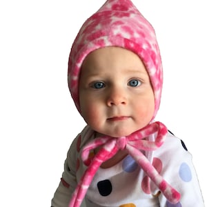 May include: A baby wearing a pink and white tie-dye hooded hat with a matching tie. The baby is wearing a white onesie with colourful polka dots.