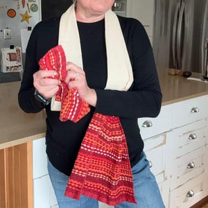 May include: A red and orange woven kitchen towel with a striped pattern is held in front of a person. A cream-colored towel is draped around the person's neck. The kitchen has a white island with silver drawer pulls.