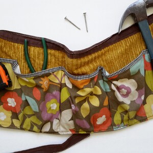 May include: A brown and yellow floral fabric tool belt with multiple pockets. The belt has a hammer, tape measure, and pliers in the pockets.