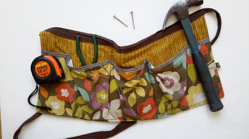 May include: A brown and yellow floral fabric tool belt with multiple pockets. The belt has a hammer, tape measure, and pliers in the pockets.