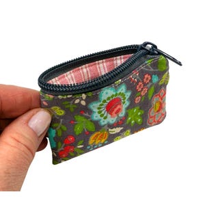 May include: A small gray zippered pouch with a floral print featuring colorful flowers and leaves. The pouch is open and the inside lining is pink and white checkered.