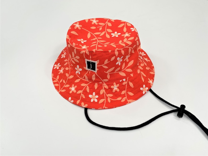 May include: A red bucket hat with a floral pattern and a black adjustable strap. The hat has a small black square label with a white letter "J" on it.