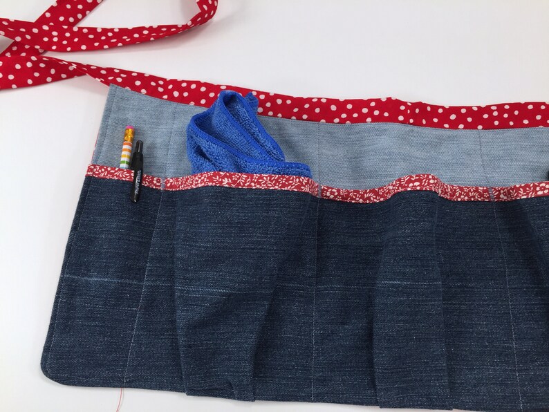 May include: A blue denim utility apron with red and white polka dot trim and multiple pockets. A blue cleaning cloth and two pencils are in the pockets.