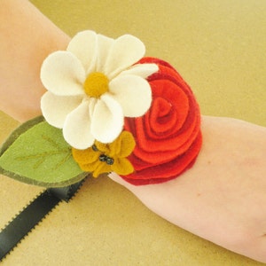 May include: A felt flower bracelet with a white daisy, a red rose, and a yellow flower with black beads. The bracelet is on a person's wrist.