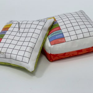 May include: Two white fabric pincushions with a grid pattern and a colour-coded chart for different sewing needle sizes. The pincushions are filled with stuffing and have a green and orange fabric trim.