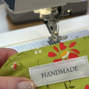 Puede incluir: Primer plano de una tela floral verde con una etiqueta blanca "HANDMADE", cosida en una máquina de coser. La tela tiene diseños de flores rojas y amarillas. La máquina de coser es blanca y gris.