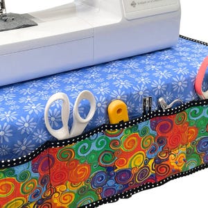 Puede incluir: Una máquina de coser se encuentra sobre una cubierta azul con estampado floral y un bolsillo colorido. El bolsillo presenta un vibrante diseño de remolino en rojo, naranja, amarillo, verde y azul. Se ven tijeras blancas, un portabobinas amarillo y otras herramientas de costura.