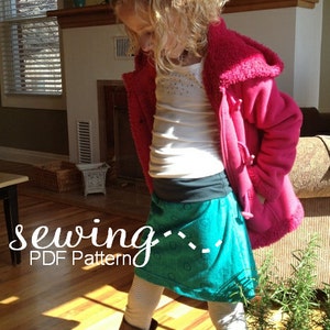 May include: A girl wearing a teal skirt with a black waistband, a white shirt, and a pink fleece jacket. The text "sewing... PDF Pattern" is on the floor in front of her.