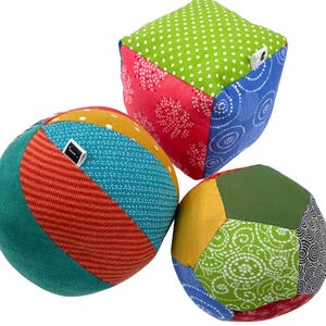 May include: Three colorful fabric toys for babies and toddlers. The toys are a ball, a cube, and a ball with multiple sides. The toys are made of different patterned fabrics in shades of red, green, blue, yellow, and orange.