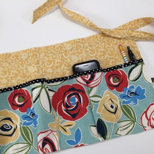 May include: A blue and yellow floral fabric utility apron with multiple pockets. The apron has a black and white polka dot trim and a yellow fabric tie.