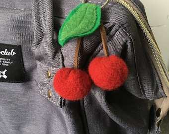 May include: A gray backpack with a cherry keychain. The keychain is made of felt and features two red cherries with green leaves.