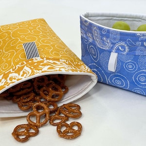 May include: Two fabric bags, one yellow with a white and black stripe, and one blue with a white paisley pattern. The yellow bag is filled with pretzels and is spilling out.