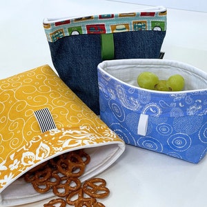 May include: Three fabric bags with different patterns. The bag on the left is yellow with a white swirl pattern and has pretzels spilling out of it. The bag in the center is blue with a white paisley pattern and has green grapes inside. The bag on the right is denim blue with a colorful patterned trim.