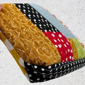 May include: A close-up of a quilted fabric pouch with a patchwork design. The pouch is made of different fabrics with polka dots, floral patterns, and solid colors. The pouch is sewn with a contrasting black thread.