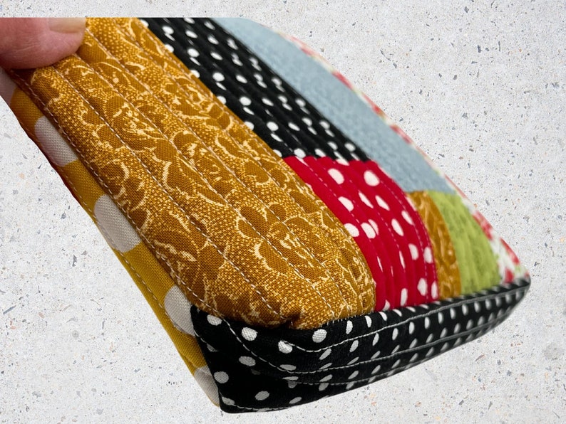May include: A close-up of a quilted fabric pouch with a patchwork design. The pouch is made of different fabrics with polka dots, floral patterns, and solid colors. The pouch is sewn with a contrasting black thread.